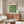 May include: Framed Bauhaus poster with green leaf pattern displayed above wooden sideboard in minimalist living room with white curved chair, glass coffee table, and sculptural table lamp. Poster features geometric green shapes arranged in repeating grid pattern on cream background with black text header.