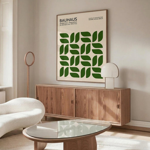 May include: Framed Bauhaus poster with green leaf pattern displayed above wooden sideboard in minimalist living room with white curved chair, glass coffee table, and sculptural table lamp. Poster features geometric green shapes arranged in repeating grid pattern on cream background with black text header.