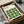 May include: Framed Bauhaus-inspired poster with green leaf pattern arranged in geometric grid on cream background, packed in cardboard shipping box with protective tissue paper and wooden corner guards, photographed on white fabric surface next to black bowl