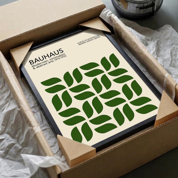 May include: Framed Bauhaus-inspired poster with green leaf pattern arranged in geometric grid on cream background, packed in cardboard shipping box with protective tissue paper and wooden corner guards, photographed on white fabric surface next to black bowl