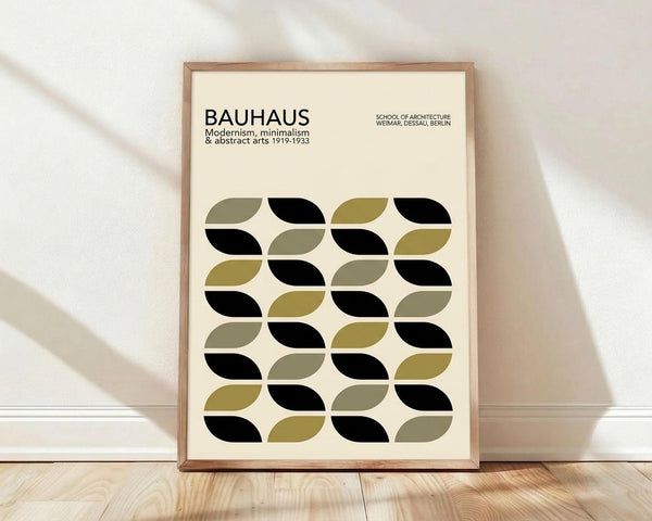 May include: Framed poster with cream background featuring geometric pattern of interlocking leaf or petal shapes in black, olive green, and muted yellow-green. Text reads "BAUHAUS Modernism, minimalism & abstract arts 1919-1933" and "SCHOOL OF ARCHITECTURE WEIMAR, DESSAU, BERLIN." Natural wood frame leans against white wall on light wooden floor with soft natural lighting casting shadows.