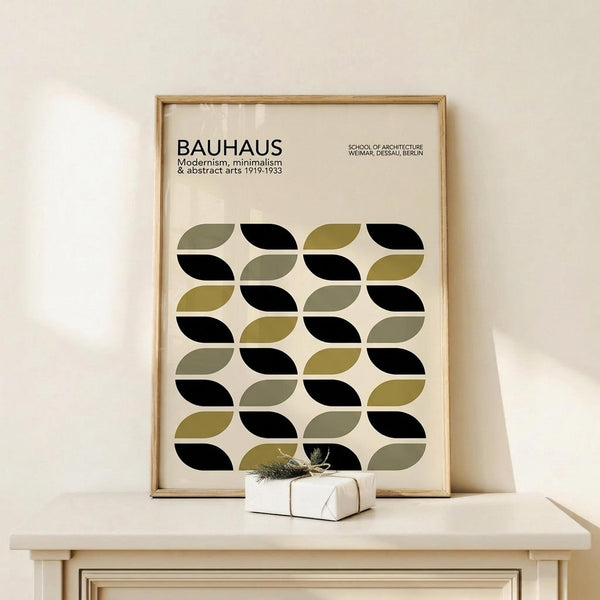May include: Framed poster displaying geometric pattern of interlocking curved shapes in black, olive green, and sage green on cream background, with "BAUHAUS" text at top. Natural wood frame sits on light wooden surface beside small wrapped gift with twine and dried botanicals against neutral wall.