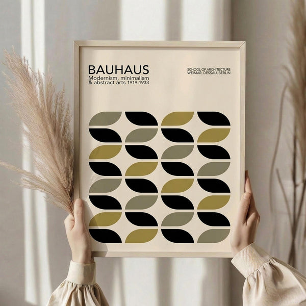 May include: Person in cream sweater holding framed poster with geometric pattern of interlocking oval shapes in black, olive green, and muted yellow on beige background. Dried pampas grass visible beside figure against soft neutral curtained backdrop.