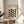 May include: Framed Bauhaus poster with geometric leaf pattern in sage green, olive, and black on cream background, displayed on round wooden side table next to beige upholstered chair. Natural lighting creates soft shadows across minimalist interior with white walls and plant leaves visible in corner.