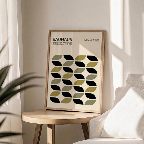 May include: Framed Bauhaus poster with geometric leaf pattern in sage green, olive, and black on cream background, displayed on round wooden side table next to beige upholstered chair. Natural lighting creates soft shadows across minimalist interior with white walls and plant leaves visible in corner.