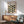 May include: Framed Bauhaus poster with geometric leaf pattern in black, olive green, and sage green displayed above a wooden sideboard. Mid-century modern living room setting features teal upholstered armchair with wood frame, white table lamp, and round coffee table. Natural sunlight creates diagonal shadows on white wall.