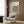 May include: Framed Bauhaus poster with geometric leaf pattern in black, olive green, and sage on cream background, displayed above a beige upholstered armchair with geometric patterned pillow. Modern living room setting features wood paneled wall, brass floor lamp with white circular base, and dark round side table with open book.