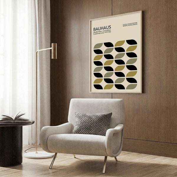 May include: Framed Bauhaus poster with geometric leaf pattern in black, olive green, and sage on cream background, displayed above a beige upholstered armchair with geometric patterned pillow. Modern living room setting features wood paneled wall, brass floor lamp with white circular base, and dark round side table with open book.