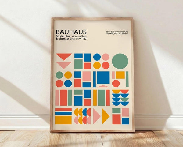 May include: Framed Bauhaus poster with colorful geometric shapes including circles, squares, triangles and rectangles in blue, red, orange, green and pink against cream background. Features "BAUHAUS" text and "School of Architecture Weimar, Dessau, Berlin" subtitle. Light wood frame sits on wooden floor against white wall with natural lighting creating soft shadows.