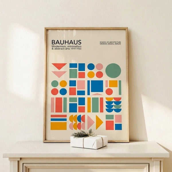 May include: Framed Bauhaus poster with colorful geometric shapes in red, blue, yellow, green, and pink arranged in grid pattern on cream background. Modern minimalist print displayed on light wood shelf with small wrapped gift box. Clean contemporary interior styling with neutral tones and natural lighting.