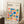 May include: Framed Bauhaus poster with colorful geometric shapes in blue, red, orange, green, and pink displayed in minimalist room setting. Poster features circles, squares, triangles and text about modernism and abstract arts 1919-1933. Light wooden frame leans against white wall near textured rug, wooden side table with white coffee cup, and beige throw blanket.
