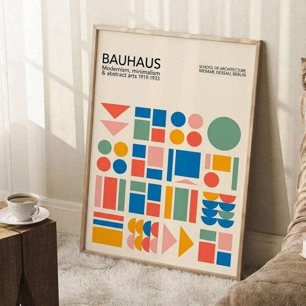 May include: Framed Bauhaus poster with colorful geometric shapes in blue, red, orange, green, and pink displayed in minimalist room setting. Poster features circles, squares, triangles and text about modernism and abstract arts 1919-1933. Light wooden frame leans against white wall near textured rug, wooden side table with white coffee cup, and beige throw blanket.
