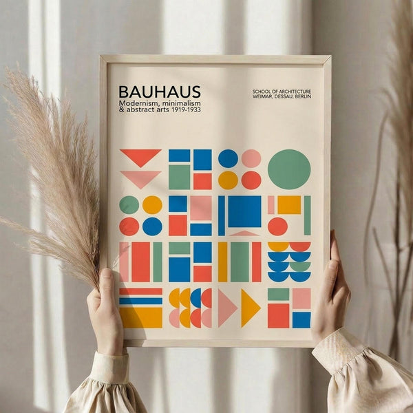 May include: Hands holding a framed Bauhaus-inspired poster with geometric shapes in blue, orange, green, pink, and red arranged in a grid pattern. The print features circles, squares, triangles, and semicircles on cream background with black text reading "BAUHAUS" and "School of Architecture Weimar, Dessau, Berlin" and dates "1919-1933". Light wood frame, neutral beige setting with dried pampas grass visible on left side.