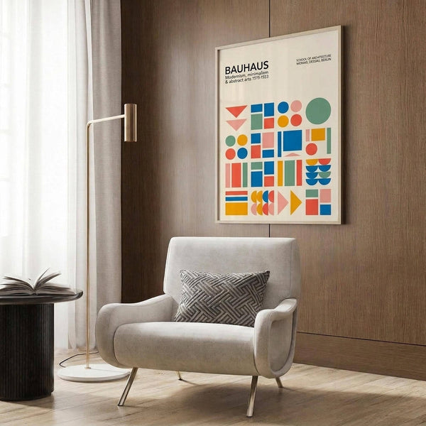 May include: Framed Bauhaus poster with colorful geometric shapes in red, blue, green, orange, and pink displayed on wood paneled wall above beige upholstered armchair with geometric patterned pillow. Modern brass floor lamp and dark round side table with open book complete the minimalist living room scene with light wood flooring.