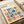 May include: Framed Bauhaus poster with colorful geometric shapes including circles, squares, triangles and rectangles in blue, red, orange, green and pink on cream background. Print sits on light wooden surface next to rolled paper tube, showing modernist design elements arranged in grid-like composition with clean typography.