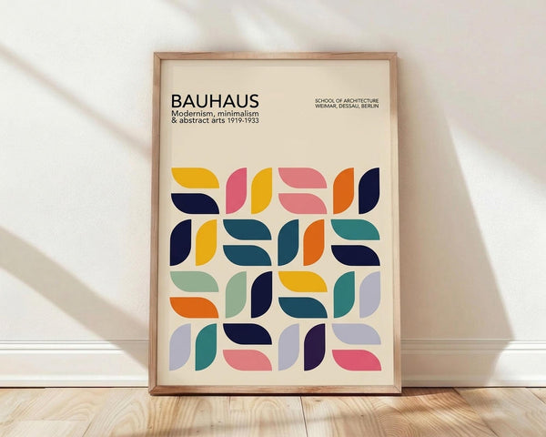May include: Framed Bauhaus poster with colorful geometric half-circle and curved shapes in yellow, pink, orange, navy, teal, and purple on cream background. Modern minimalist design leaning against white wall on light wood floor with soft natural lighting and shadows.