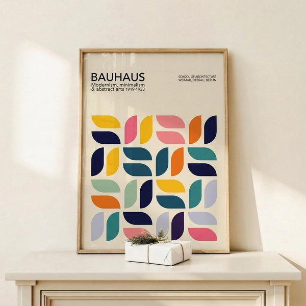 May include: Framed Bauhaus poster with geometric pattern of colorful quarter-circle shapes in yellow, pink, blue, orange, and teal, displayed on light wood surface next to small wrapped gift with evergreen sprig against cream wall