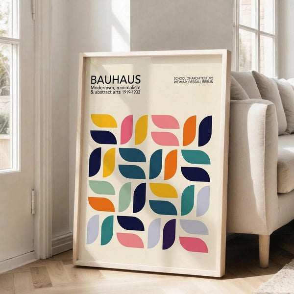 May include: Framed Bauhaus-inspired poster with colorful geometric shapes in a grid pattern, featuring semicircles and quarter-circles in navy, orange, pink, teal, yellow, and purple on cream background. Shows black text reading "BAUHAUS Modernism, minimalism & abstract arts 1919-1933" and "School of Architecture Weimar, Dessau, Berlin." Light wood frame leaning against white wall next to beige sofa in bright, modern living room with hardwood floors.