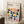 May include: Framed Bauhaus poster with colorful geometric leaf-like shapes in a grid pattern, featuring navy, yellow, pink, orange, teal, and purple elements on cream background. Light wood frame leans against white wall corner next to textured throw blanket and wooden block with white coffee cup and saucer.