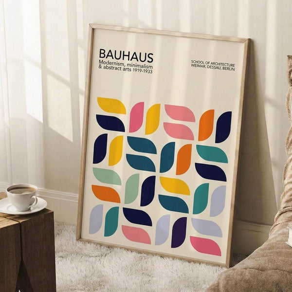 May include: Framed Bauhaus poster with colorful geometric leaf-like shapes in a grid pattern, featuring navy, yellow, pink, orange, teal, and purple elements on cream background. Light wood frame leans against white wall corner next to textured throw blanket and wooden block with white coffee cup and saucer.
