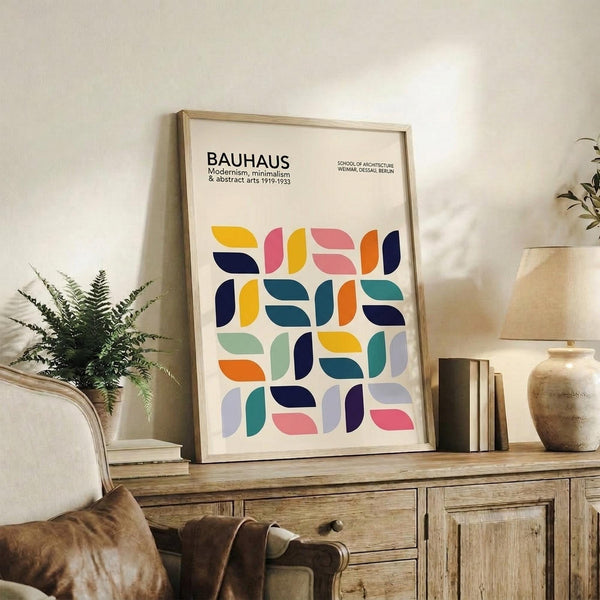 May include: Framed Bauhaus poster with colorful geometric semicircle pattern in yellow, pink, orange, navy, teal and green, displayed on rustic wooden sideboard next to fern plant, books, and ceramic vases in neutral living room setting with beige chair visible in corner.
