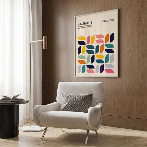 May include: Framed Bauhaus poster with colorful geometric leaf-like shapes in yellow, pink, orange, teal, and navy blue displayed on wood-paneled wall above light gray upholstered armchair with patterned pillow, next to brass floor lamp and dark round side table with open book