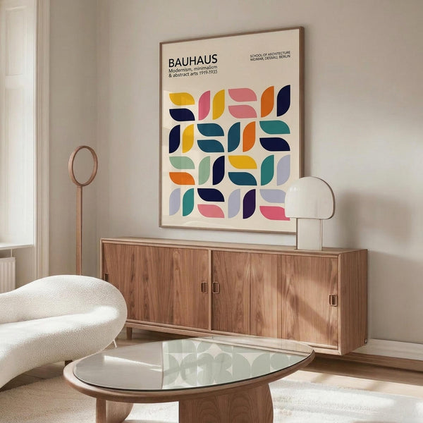 May include: Framed Bauhaus poster with colorful geometric half-circle pattern in yellow, pink, orange, navy, and teal displayed above wooden sideboard. Modern living room setting with white curved chair, glass oval coffee table, and brass floor lamp. Clean minimalist interior with white walls and natural lighting.