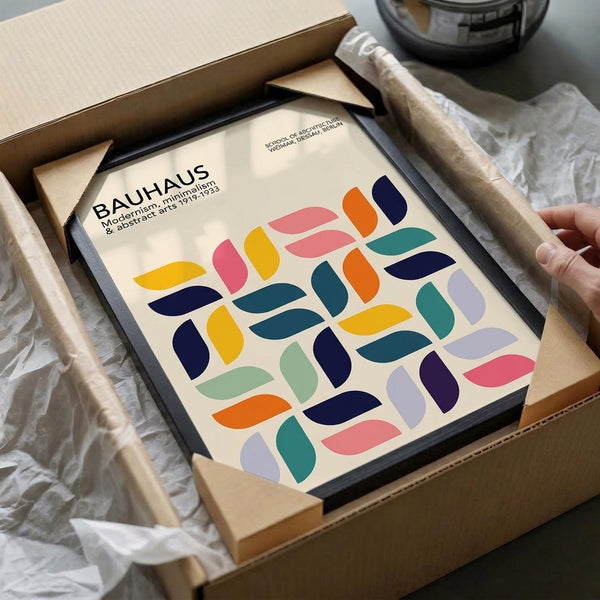 May include: Framed Bauhaus poster with colorful geometric leaf pattern in pink, blue, yellow, orange, and green shapes on cream background, shown being unboxed from cardboard packaging with protective wrapping and wooden frame corners visible on white surface.