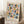 May include: Framed geometric art print with colorful semicircles and half-moons in muted pink, blue, green, yellow, and navy arranged in a grid pattern on cream background. Light wood frame leans against white wall corner next to wooden side table with white coffee cup and saucer, beige knit throw blanket visible on textured white rug.