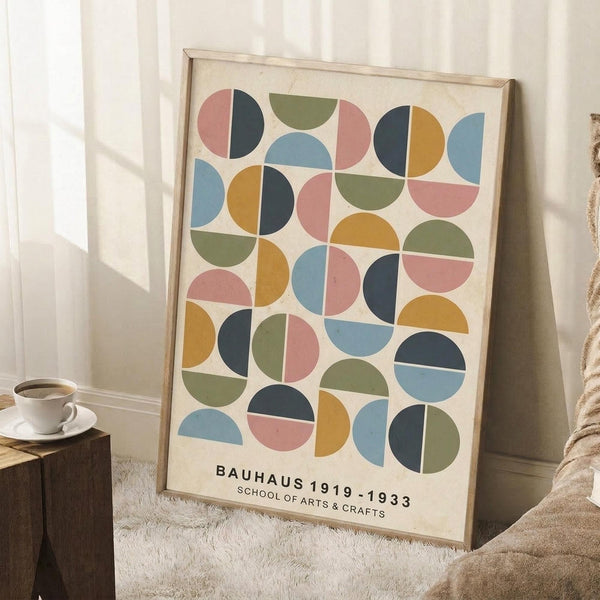 May include: Framed geometric art print with colorful semicircles and half-moons in muted pink, blue, green, yellow, and navy arranged in a grid pattern on cream background. Light wood frame leans against white wall corner next to wooden side table with white coffee cup and saucer, beige knit throw blanket visible on textured white rug.