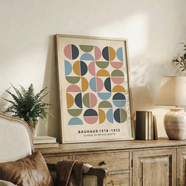 May include: Framed poster with colorful geometric circles and semicircles in muted pink, blue, green, orange and navy tones on cream background, displayed on rustic wood sideboard alongside fern plant, ceramic vase, and stacked books in cozy living room setting with beige upholstered furniture and soft natural lighting.