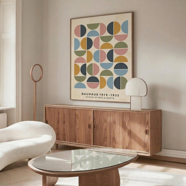 May include: Modern living room interior with geometric poster on wall featuring colorful circles and semicircles in pink, blue, green, orange, and black on cream background. Walnut credenza below with white mushroom lamp and copper floor lamp. White curved chair and glass-top oval coffee table complete the mid-century modern setting with neutral walls.