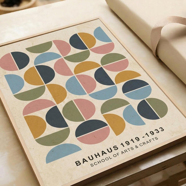 Modernist geometric poster featuring colorful circles divided into half and quarter sections in muted tones of sage green, dusty pink, navy blue, mustard yellow, and light blue on cream background. Text reads "BAUHAUS 1919-1933 SCHOOL OF ARTS & CRAFTS" in black lettering. Print lies flat on wooden surface next to rolled paper tubes, showing natural paper texture and clean graphic design style.