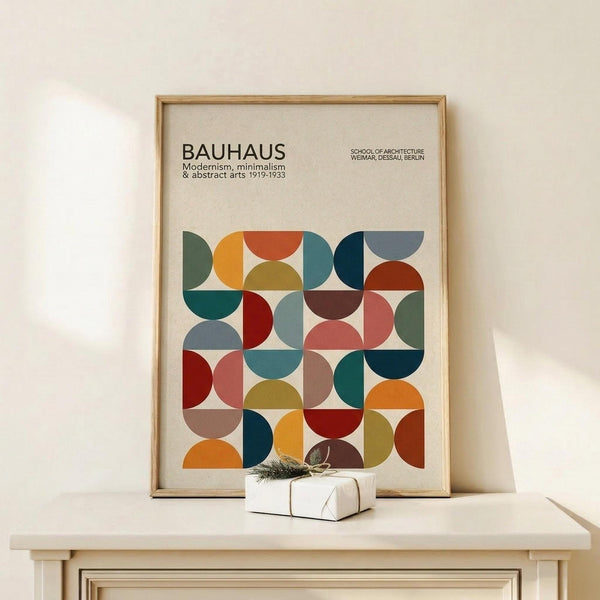 May include: Framed Bauhaus poster with geometric pattern of colorful overlapping semicircles in orange, teal, red, brown, and muted tones, displayed on light wood console table next to small wrapped gift with evergreen sprig, against cream wall with natural lighting