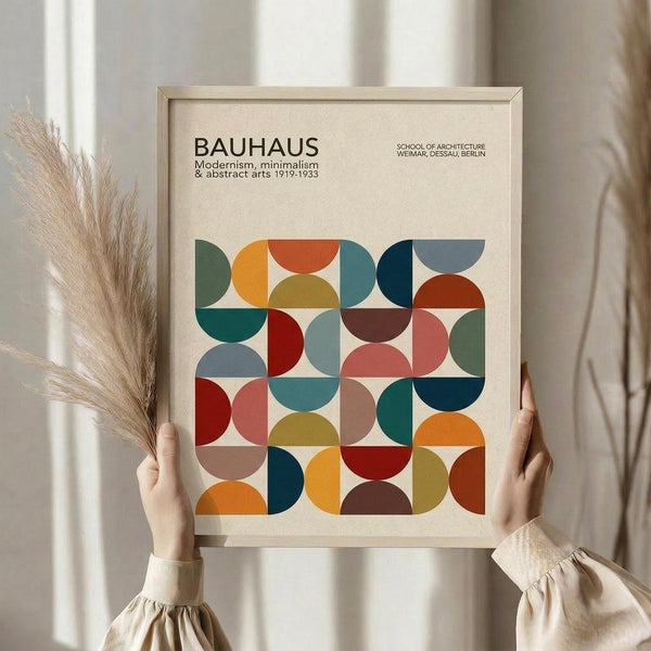 May include: Hands holding a framed geometric art print with colorful overlapping circles and semicircles in orange, teal, red, brown, and sage green on cream background. Light wooden frame, dried pampas grass visible to left, neutral beige clothing and curtained window setting.