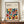 May include: Framed poster with geometric pattern of colorful semicircles and quarter-circles in squares, featuring text about Bauhaus modernism and minimalism. Earth-tone and jewel-tone colors including orange, teal, red, olive, and burgundy on cream background. Brown wooden frame displayed on walnut credenza next to white ceramic vase and stacked books in bright room with white paneled walls.