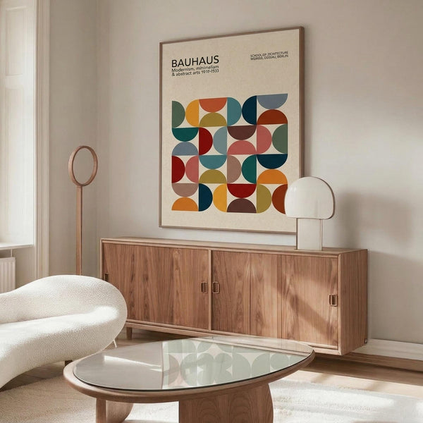 May include: Framed Bauhaus poster with colorful geometric circles and semicircles in red, blue, green, orange, and yellow arranged in a grid pattern, displayed above a mid-century walnut credenza with sliding doors. White modern chair and glass-top oval coffee table visible in minimalist living room setting with white walls and natural lighting.
