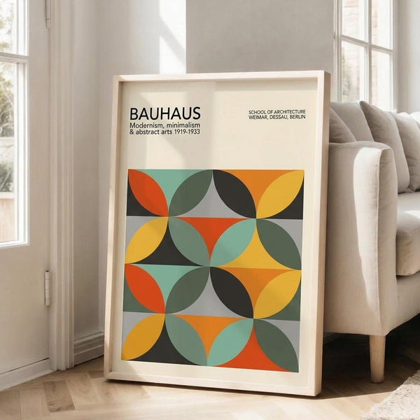 May include: Framed poster with geometric Bauhaus-inspired design featuring overlapping circles in orange, yellow, teal, olive green, and black creating a repeating pattern. Light wood frame leaning against white wall next to beige upholstered chair in bright, naturally-lit room with hardwood floors and large windows.