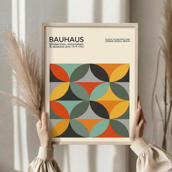 May include: Person holding framed geometric poster with colorful overlapping circles in orange, yellow, teal, and dark green creating diamond patterns. Light wood frame against neutral curtain backdrop with dried pampas grass visible on left side.