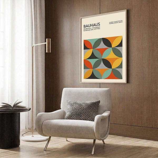 May include: Framed Bauhaus poster with colorful geometric circles in orange, yellow, green, and red displayed on wood-paneled wall above beige upholstered armchair with patterned throw pillow. Modern brass floor lamp and round black side table with open book complete the minimalist living room setting with light wood floors.