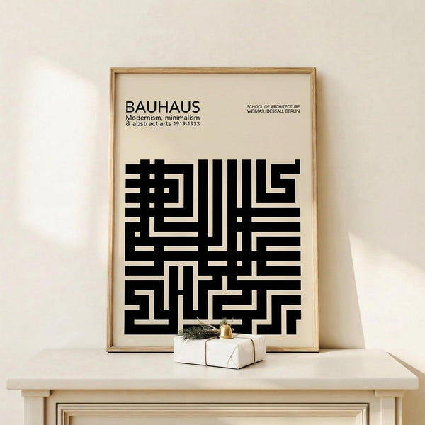 May include: Framed Bauhaus poster with black geometric maze-like pattern on cream background, displayed on light wood console table against beige wall. Small white gift box with greenery and gold bell sits in front of frame. Natural wood frame matches minimalist Scandinavian-style furniture. Soft natural lighting creates gentle shadows on textured wall surface.