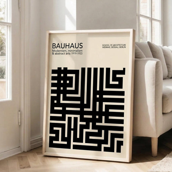 May include: Black and cream geometric abstract poster in light wood frame leaning against white wall next to beige upholstered chair. Design features bold rectangular maze-like pattern with "BAUHAUS" text and "Modernism, minimalism & abstract arts 1919-1933" subtitle. Natural lighting creates soft shadows on hardwood floor in minimalist room setting.