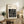 May include: Framed Bauhaus poster with black geometric maze-like pattern on cream background, displayed on rustic wooden sideboard alongside fern plant, stacked books, ceramic vase, and brown leather chair in warm-toned living room setting