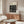 May include: Black and white geometric Bauhaus poster with abstract maze-like pattern mounted above wooden credenza in modern living room. Cream colored sculptural chair and glass coffee table visible. Minimalist interior with neutral tones and natural lighting from window.