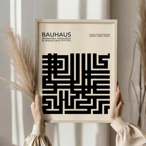 May include: Person in cream sweater holding framed Bauhaus poster against white curtained window, with dried pampas grass beside them. Black geometric maze-like design on cream background with "BAUHAUS" text and "Modernism, minimalism & abstract arts 1919-1933" subtitle. Light wood frame complements neutral aesthetic setting.