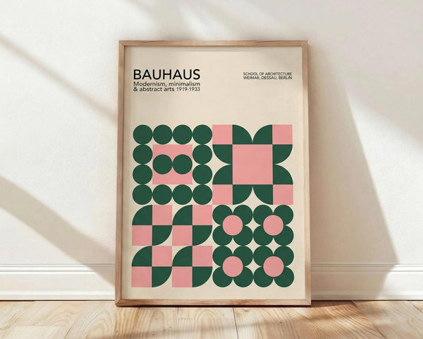 May include: Framed Bauhaus modernist poster with geometric design featuring green circles and pink squares arranged in abstract patterns. Cream background with black text reading "BAUHAUS Modernism, minimalism & abstract arts 1919-1933" and "SCHOOL OF ARCHITECTURE WEIMAR, DESSAU, BERLIN." Light wood frame displayed against white wall on wooden floor with natural lighting creating soft shadows.