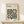 May include: Framed Bauhaus-style poster with geometric pattern in forest green and coral pink circles and squares, displayed on light wood console table beside white wrapped gift with twine and evergreen sprig against cream wall