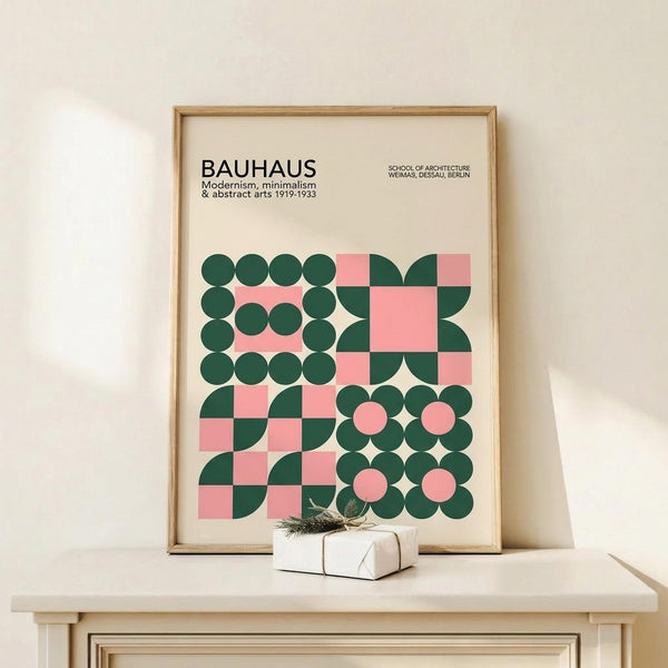 May include: Framed Bauhaus-style poster with geometric pattern in forest green and coral pink circles and squares, displayed on light wood console table beside white wrapped gift with twine and evergreen sprig against cream wall