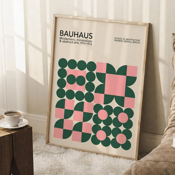 May include: A framed Bauhaus poster with geometric green circles and pink squares on cream background, leaning against white wall beside wooden cube table with white coffee cup and beige textured throw blanket on white fluffy rug.