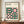 May include: Framed Bauhaus-style poster with geometric design featuring pink and dark green circles, quarter-circles, and squares arranged in a grid pattern. Text reads "BAUHAUS Modernism, minimalism & abstract arts 1919-1933" and "SCHOOL OF ARCHITECTURE WEIMAR, DESSAU, BERLIN." Brown wooden frame displayed on wooden sideboard alongside white ceramic vase and stacked books in elegant room with cream walls and white molding.